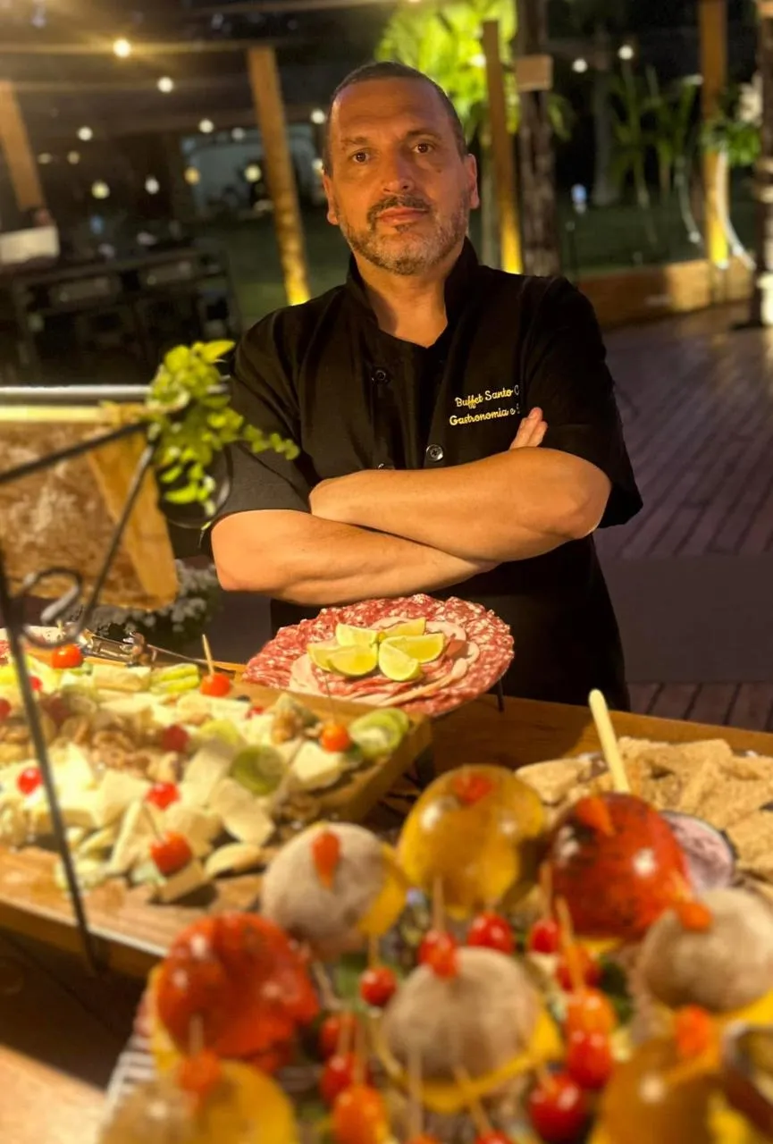 Chef do D’Faria à frente da ilha de frios, com prato de salame fatiado com limão e tábuas de queijos, frios e espetinhos de frutas